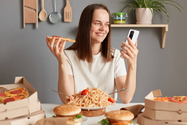 Photo of cheerful Caucasian young adult woman with brown hair wearing white t shirt sitting in kitchen at table and looking at smart phone display, having delicious snack, eating pizza. - Фото, зображення