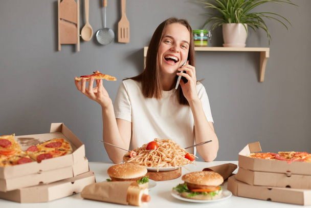 Photo of joyful cheerful attractive woman with brown hair wearing white t shirt sitting in kitchen at table and talking on smart phone, laughing happily, having dinner, eating pizza. - Фото, зображення