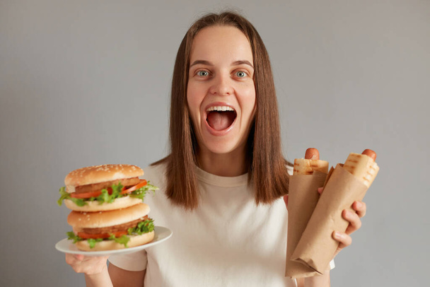 Portrait of extremely happy woman eating fast food, holding hot dog and sandwich wearing white T-shirt posing isolated over gray background, rejoicing to have cheat meal. - Photo, Image