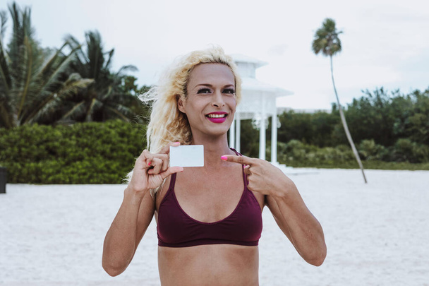 Young transgender man wearing makeup and woman clothes holding a blank card with copy space at the beach in Mexico Latin America - Photo, Image