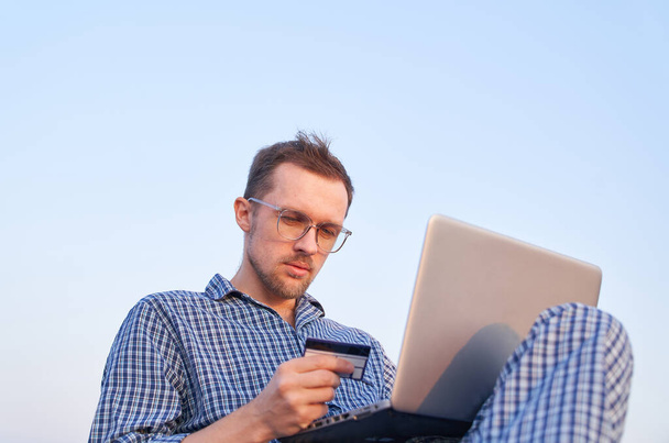 Roztomilý běloch používající notebook pro nakupování s kreditní kartou v ruce. Veselý muž v brýlích a oblečení na spaní pomocí notebooku venku v dopoledních hodinách nákupy on-line - Fotografie, Obrázek
