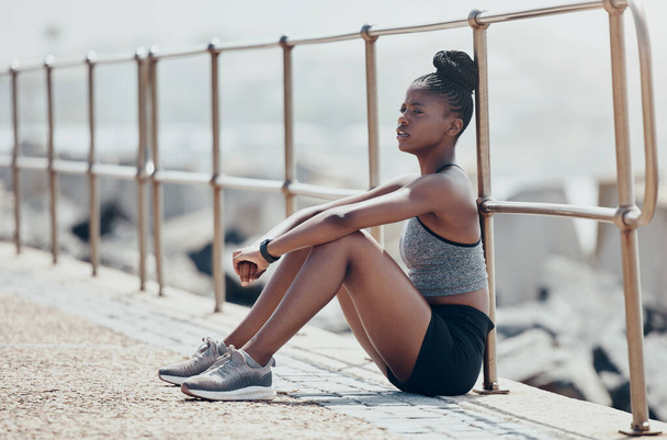 Strand, pihenés és női futó gondolkodás után egy szabadtéri kardió edzés a természetben az óceán. Fáradt afrikai sportoló, futó és fitness lány ül a sétányon lélegezni, nyugodt és pihenni edzés után. - Fotó, kép