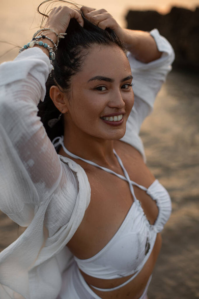 Portrait of young tanned sexy woman in white swimsuit with her hands raised up with smile looking at amera against the background of ocean. Wonderful moments. Playful mood. Sunset on sea. - Photo, image