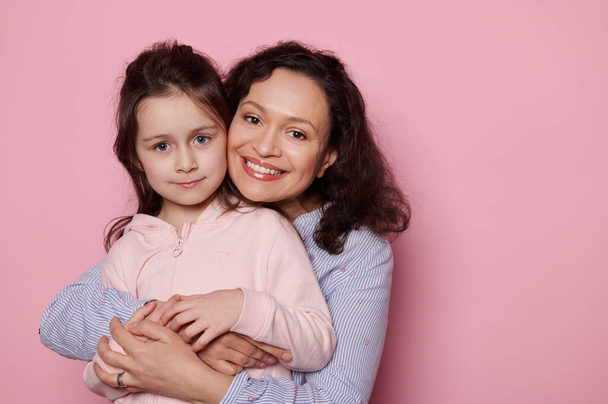 Multi-ethnic pretty woman, loving mother smiles a cheerful toothy smile looking at camera, experiencing happiness while gently hugging her adorable beloved little child girl, isolated pink background - Foto, Bild