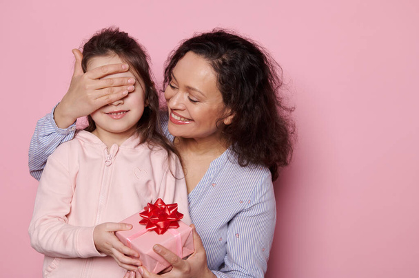 Ευτυχισμένη γυναίκα, στοργική μητέρα που κλείνει τα μάτια της υπέροχης κόρης της ενώ της δίνει ένα χαριτωμένο δώρο σε απομονωμένο ροζ φόντο. Διεθνής Ημέρα Προστασίας των Παιδιών. Ανθρώπους. Έννοια οικογενειακών σχέσεων - Φωτογραφία, εικόνα