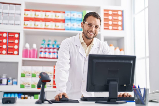 Nuori mies farmaseutti hymyilee luottavainen tietokoneella apteekissa - Valokuva, kuva