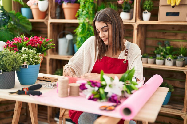 Giovane bella donna ispanica fiorista sorridente scrittura fiduciosa sul taccuino al negozio di fiori - Foto, immagini