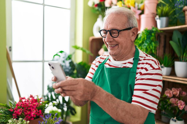 Середнього віку сіро-волохатий чоловік флорист посміхається впевнено використовуючи смартфон у квітковому магазині
 - Фото, зображення
