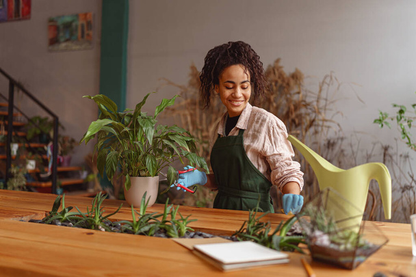 Усміхнена жінка-флорист доглядає за рослиною, поливаючи її у квітковому магазині. Високоякісна фотографія
 - Фото, зображення