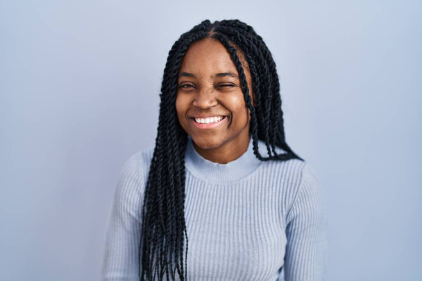 Afroamerikanerin steht vor blauem Hintergrund und zwinkert in die Kamera mit sexy Ausdruck, fröhlichem und glücklichem Gesicht.  - Foto, Bild