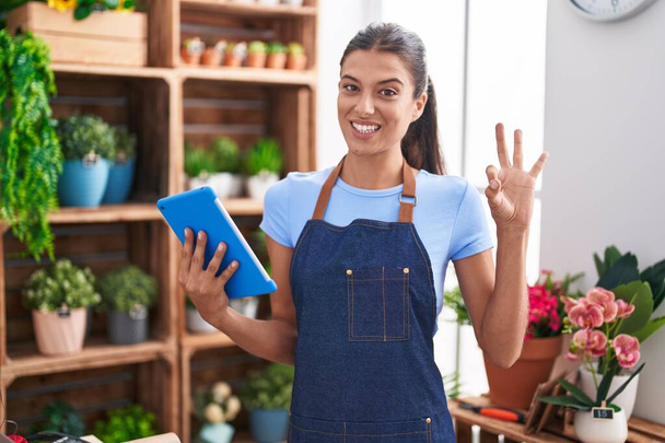 Brunette νεαρή γυναίκα που εργάζονται σε ανθοπωλείο κρατώντας tablet κάνει ok υπογράψει με τα δάχτυλα, χαμογελώντας φιλικό gesturing εξαιρετικό σύμβολο  - Φωτογραφία, εικόνα