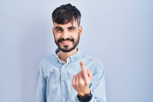 Молодой латиноамериканец с бородой стоит на синем фоне и манит сюда жест с рукой, приглашая приветствовать счастливый и улыбающийся  - Фото, изображение