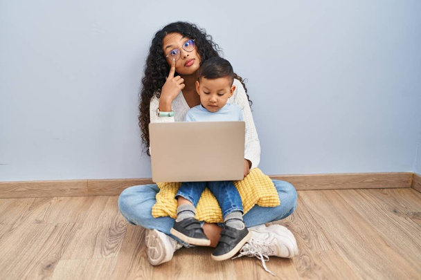 Joven madre y niño hispano usando computadora portátil sentada en el suelo apuntando al ojo observándote gesto, expresión sospechosa  - Foto, imagen