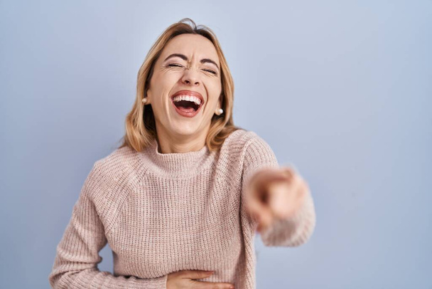Hispanic woman standing over blue background laughing at you, pointing finger to the camera with hand over body, shame expression  - Foto, immagini