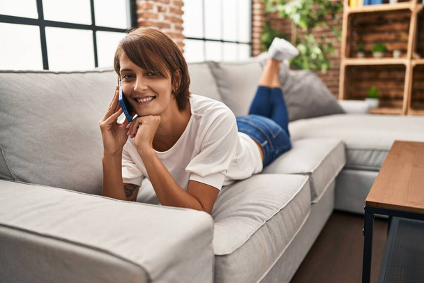 Jeune belle femme hispanique parlant sur le smartphone couché sur le canapé à la maison - Photo, image