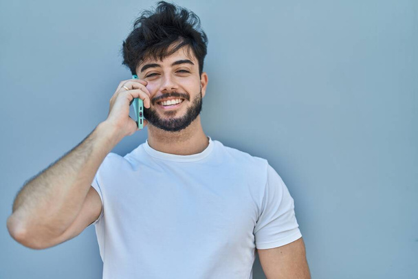 Genç İspanyol adam gülümsüyor. Akıllı telefondan beyaz izole edilmiş arka planda konuşuyor. - Fotoğraf, Görsel
