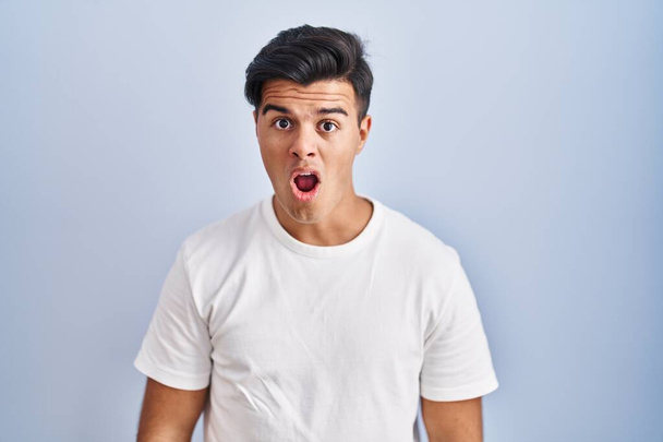 Hispanic man standing over blue background in shock face, looking skeptical and sarcastic, surprised with open mouth  - Photo, image