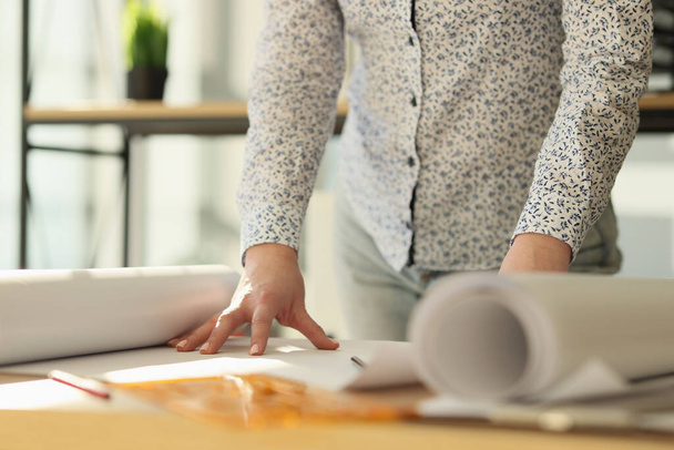 Женщина-дизайнер стоит рядом с рабочим столом с рулонами бумаги. Архитектор работает в своем офисе с крупным планом. - Фото, изображение
