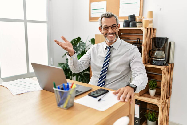 Średni wiek siwowłosy mężczyzna pracownik biznesu za pomocą laptopa mówi w biurze - Zdjęcie, obraz