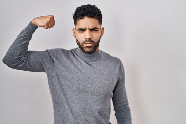 Hombre hispano con barba de pie sobre fondo blanco persona fuerte mostrando el músculo del brazo, confiado y orgulloso de poder  - Foto, Imagen