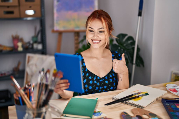Nuori valkoihoinen nainen istuu taidestudiolla tekemässä videopuhelua tabletin hymyillen onnellisena ja positiivisena, peukalo pystyssä tekemässä erinomaista ja hyväksyntämerkkiä  - Valokuva, kuva