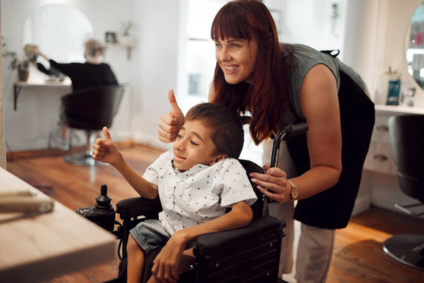 Kuaför, kuaför ve tekerlekli sandalyedeki çocuk saç kesiminden sonra başparmakları havada. Genç engelli çocuk için kuaförlük, berberlik ve saç bakımı. Destekleyin, gülümseyin ve mutlu özel ihtiyaçları olan çocuklardan teşekkür edin.. - Fotoğraf, Görsel