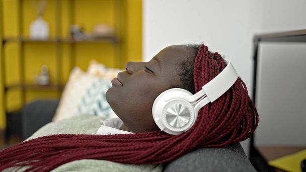 African woman with braided hair listening to music lying on sofa at home - Foto, Imagen