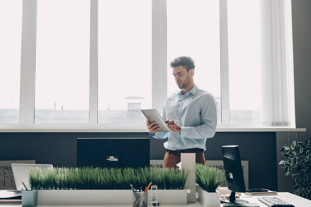 Уверенный молодой бизнесмен с помощью цифровой планшет, стоя у окна в офисе - Фото, изображение
