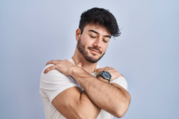 Hispanic man with beard standing over white background hugging oneself happy and positive, smiling confident. self love and self care  - Foto, immagini