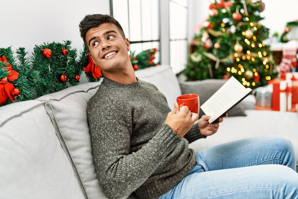 Genç İspanyol adam kitap okuyor. Evdeki Noel ağacının yanında kanepede kahve içiyor. - Fotoğraf, Görsel
