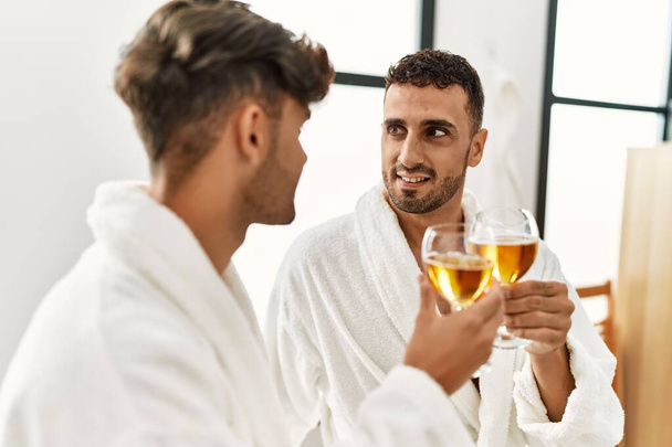 Two hispanic men couple toasting with champagne sitting on massage table at beauty center - Foto, immagini