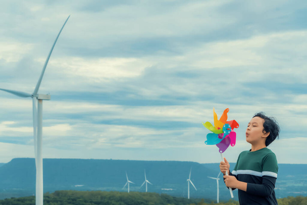 Progressive young asian boy playing with wind pinwheel toy in the wind turbine farm, green field over the hill. Green energy from renewable electric wind generator. Windmill in the countryside concept - Φωτογραφία, εικόνα