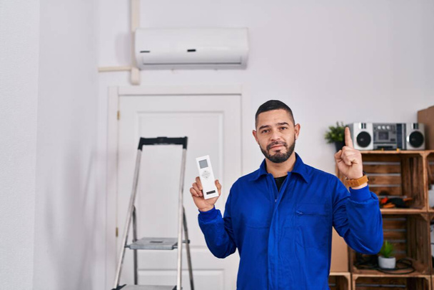 Réparateur hispanique travaillant avec climatiseur surpris par une idée ou une question pointant du doigt avec un visage heureux, numéro un  - Photo, image