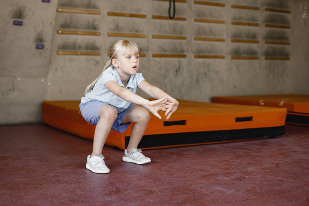 Little girl in rock-climbing gym. Girl is warming up wearing sports outfit. Blonde girl wearing shorts and t-shirt. - Fotoğraf, Görsel