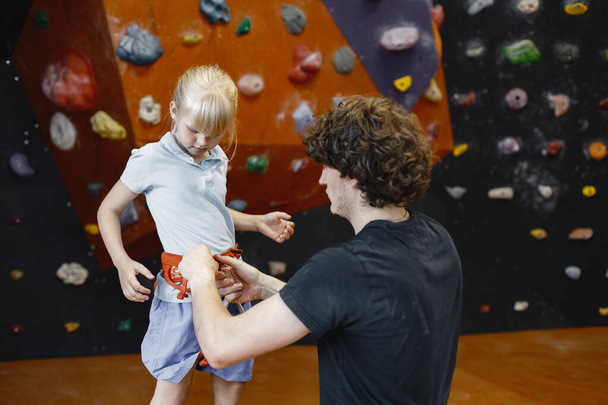 Instructor man is helping client small girl to put on safety harnesses in rock climbing center indoors. Little girl is engaged in rock climbing in the childrens entertainment center. Blonde caucasian - Fotografie, Obrázek
