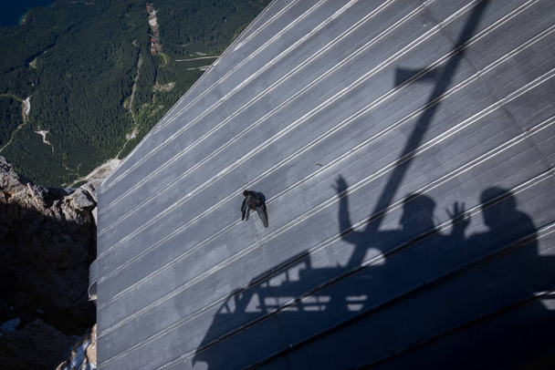 The bird sitting calmly on the roof of the building, the shadow of a man with his arms raised. Zugspitze massif in the bavarian alps - Photo, Image
