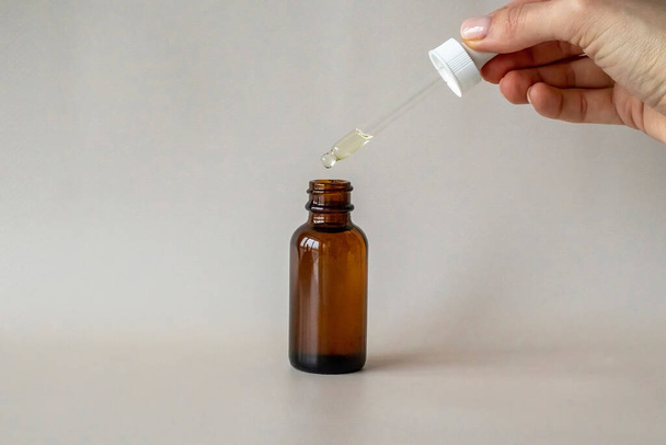 The Young Womans hand holds a bottle with a dropper, with an oil or serum for facial and body care. Picks up serum with a pipette and drops it into a brown glass bottle - Foto, Imagem