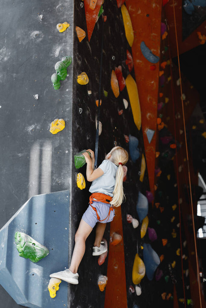Little girl in rock-climbing gym. Girl is climbing up artificial wall wearing sports outfit and safety equipment. Blonde girl wearing shorts and t-shirt. - Valokuva, kuva