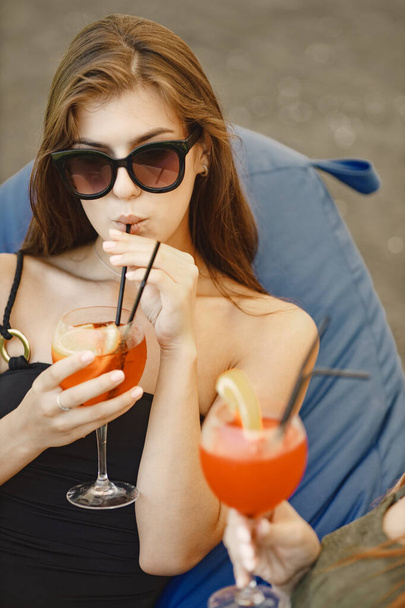Caucasian girl in swimwear and sunglasses is drinking cocktails, talking with her friend and smiling. Female friends sunbathing on the bean bag chairs on a grass. Girls wearing black and khaki - Fotoğraf, Görsel