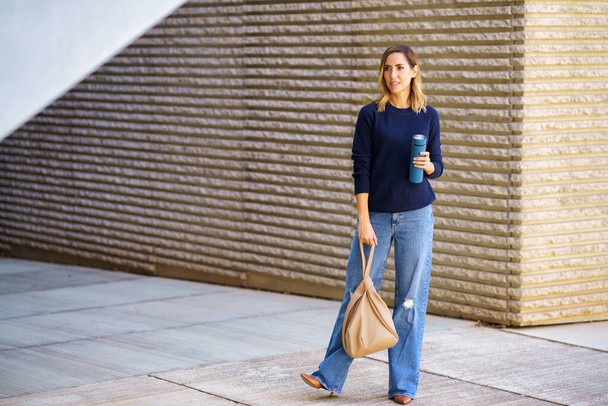 Pieno corpo donna in jeans alla moda e maglione con borsa e thermos guardando altrove mentre in piedi sul marciapiede fuori edificio moderno - Foto, immagini