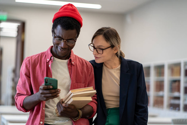 Two diverse students using mobile phone scrolling social media during study break, standing in classroom, study buddies Caucasian girl and African guy resting after studying together in library - Foto, immagini