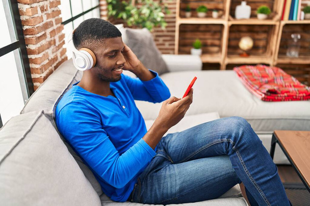 Молодой африканский американец слушает музыку, сидя дома на диване - Фото, изображение