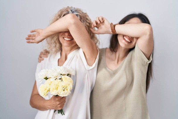 Madre e hija sosteniendo ramo de flores blancas sonriendo alegre jugando a echar un vistazo a un boo con las manos mostrando la cara. sorprendido y salido  - Foto, imagen