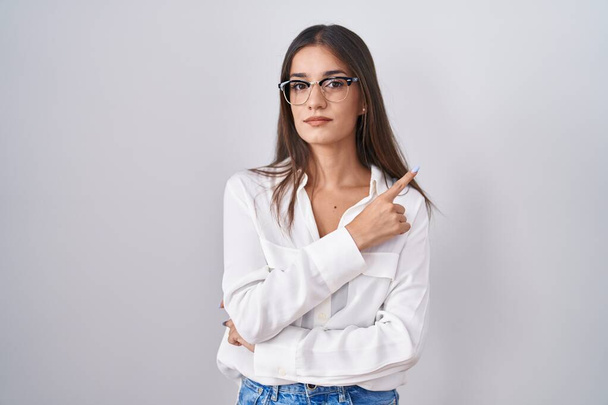 Mladá bruneta žena nosí brýle ukazující ukazováčkem na stranu zobrazující reklamu, vážnou a klidnou tvář  - Fotografie, Obrázek