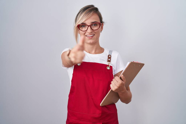 Młoda blondynka w mundurze kelnera trzymająca notatnik wskazujący palcem w kamerę ze szczęśliwą i zabawną twarzą. dobra energia i wibracje.  - Zdjęcie, obraz