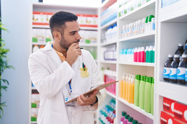 Young hispanic man pharmacist writing on document with serious expression at pharmacy - Photo, Image
