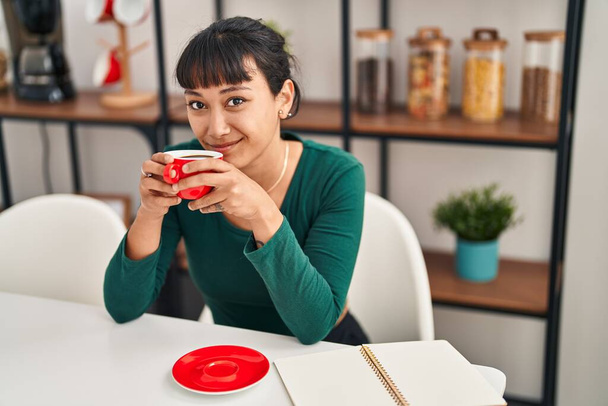 Young beautiful hispanic woman drinking coffee sitting on table at home - Photo, image