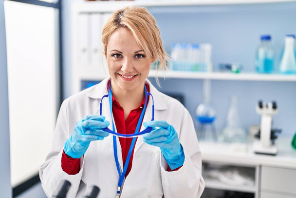 Joven rubia científica sonriendo confiada sosteniendo gafas en el laboratorio - Foto, Imagen