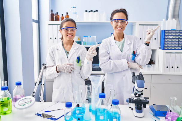 Joven madre e hija en el laboratorio científico sonriendo feliz señalando con la mano y el dedo hacia un lado  - Foto, imagen