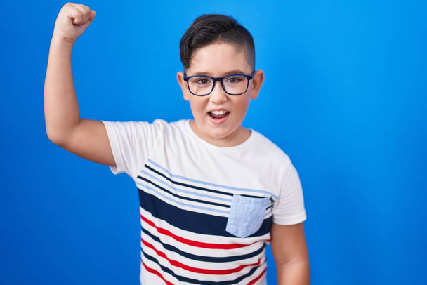 Young hispanic kid standing over blue background angry and mad raising fist frustrated and furious while shouting with anger. rage and aggressive concept.  - Fotografie, Obrázek